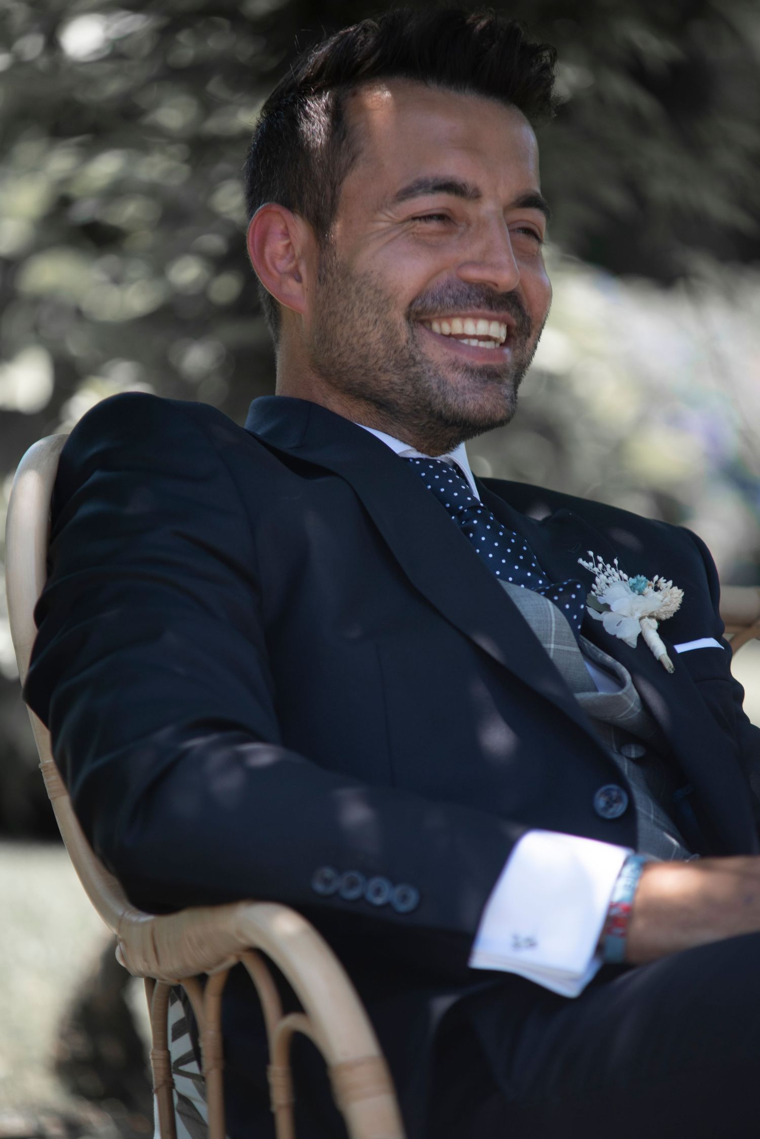 A groom in a dark suit and polka dot tie smiles while relaxing in a wooden chair outdoors.