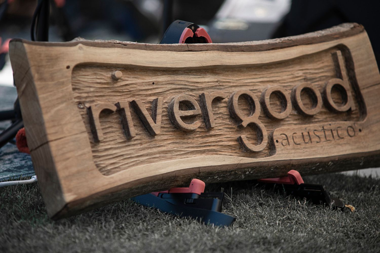 A wooden carved sign reading 'rivergood' with rustic woodgrain texture displayed on grass.