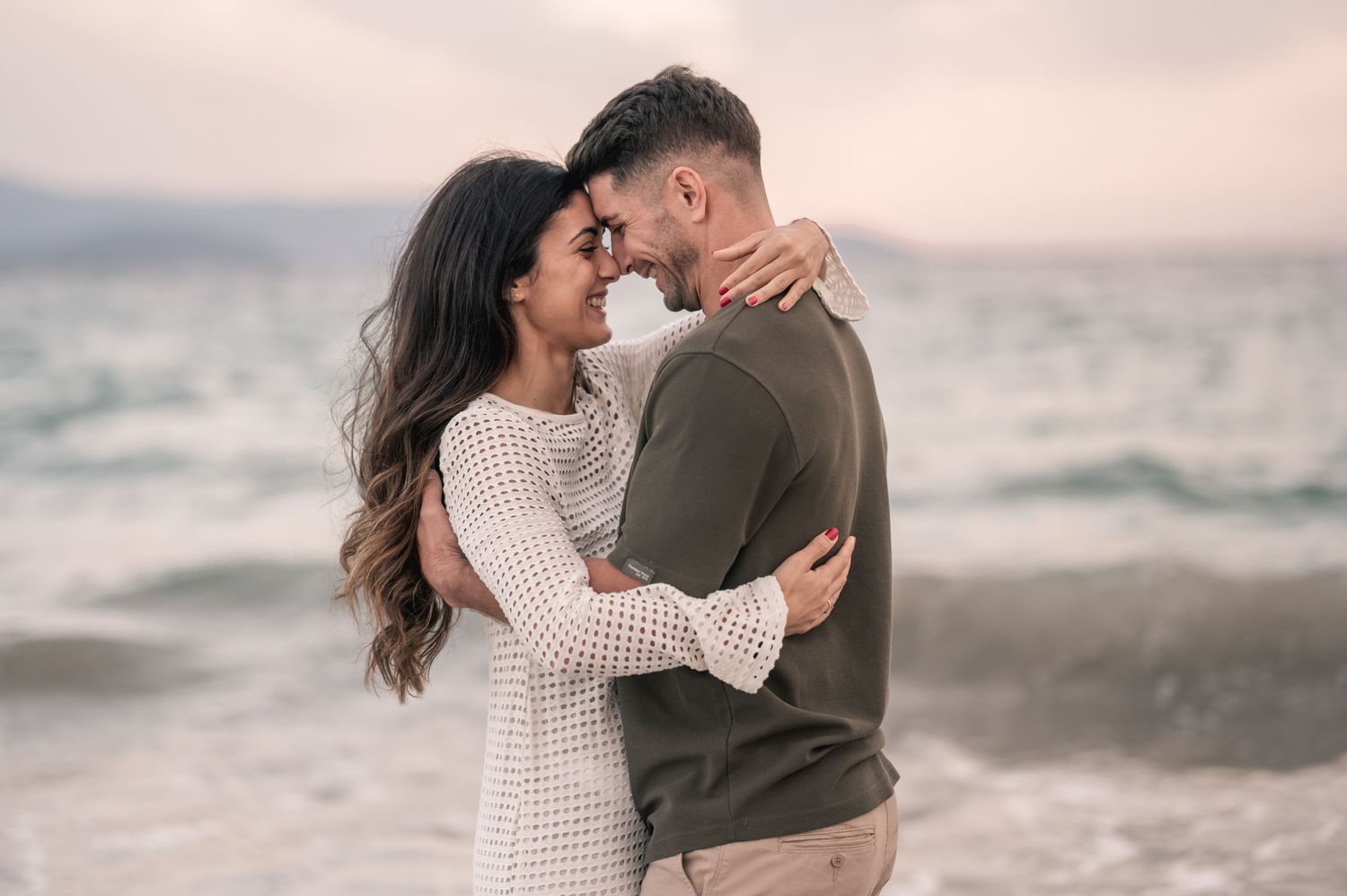 A couple in casual attire shares a tender embrace against a scenic ocean backdrop at sunset.