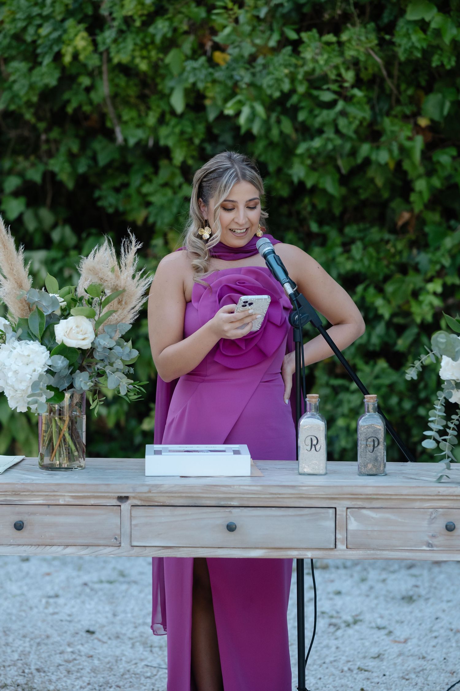 Wedding guest in purple dress smells flowers in glass vase on rustic wooden table at outdoor reception.