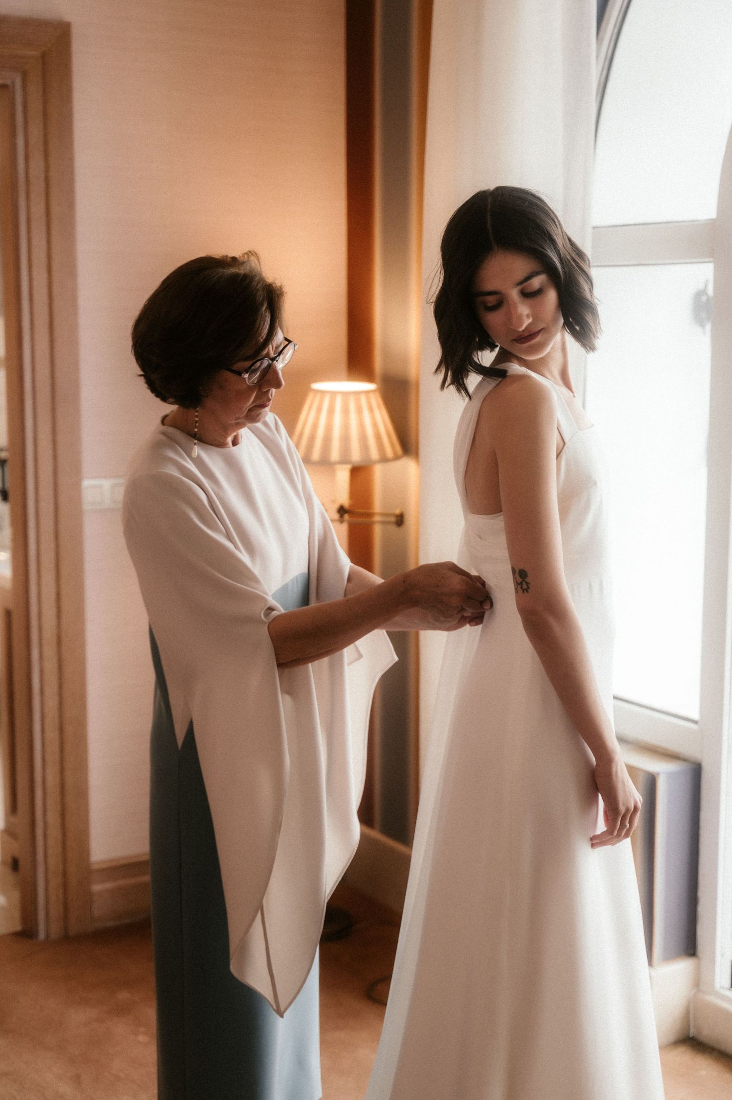 A series of intimate moments showing final adjustments being made to a white wedding dress in a sunlit room.