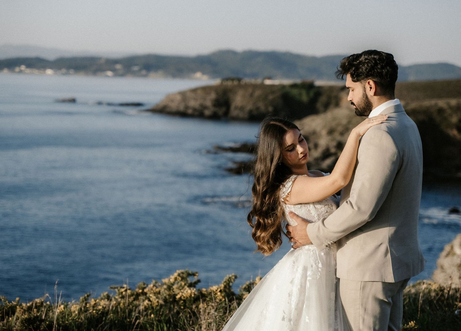 Newlyweds share an intimate moment on coastal cliffs overlooking the ocean in formal wedding attire.