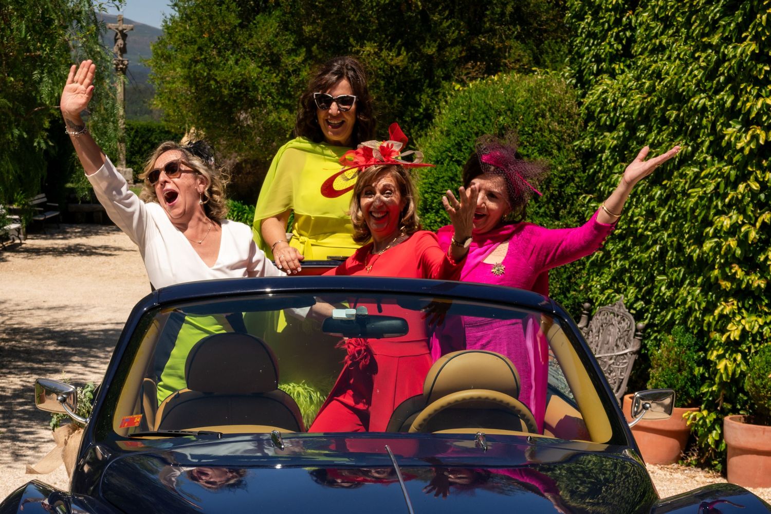 A group of people pose playfully in a red convertible car surrounded by lush green foliage on a sunny day.