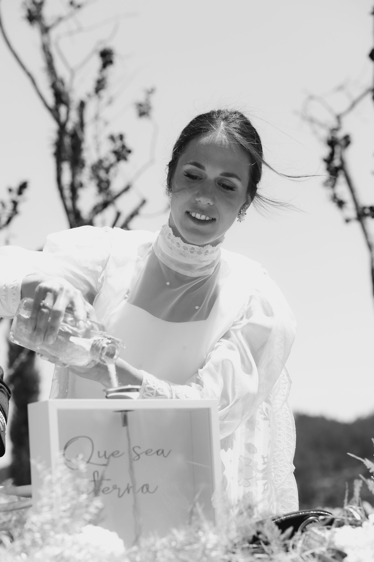 A woman in white pours liquid into a decorative box during an outdoor ceremony.