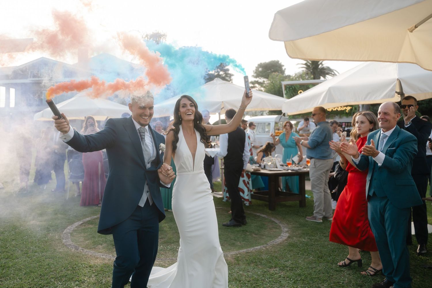 Wedding guests dance and celebrate under white tents with colorful smoke bombs creating a vibrant outdoor party atmosphere.