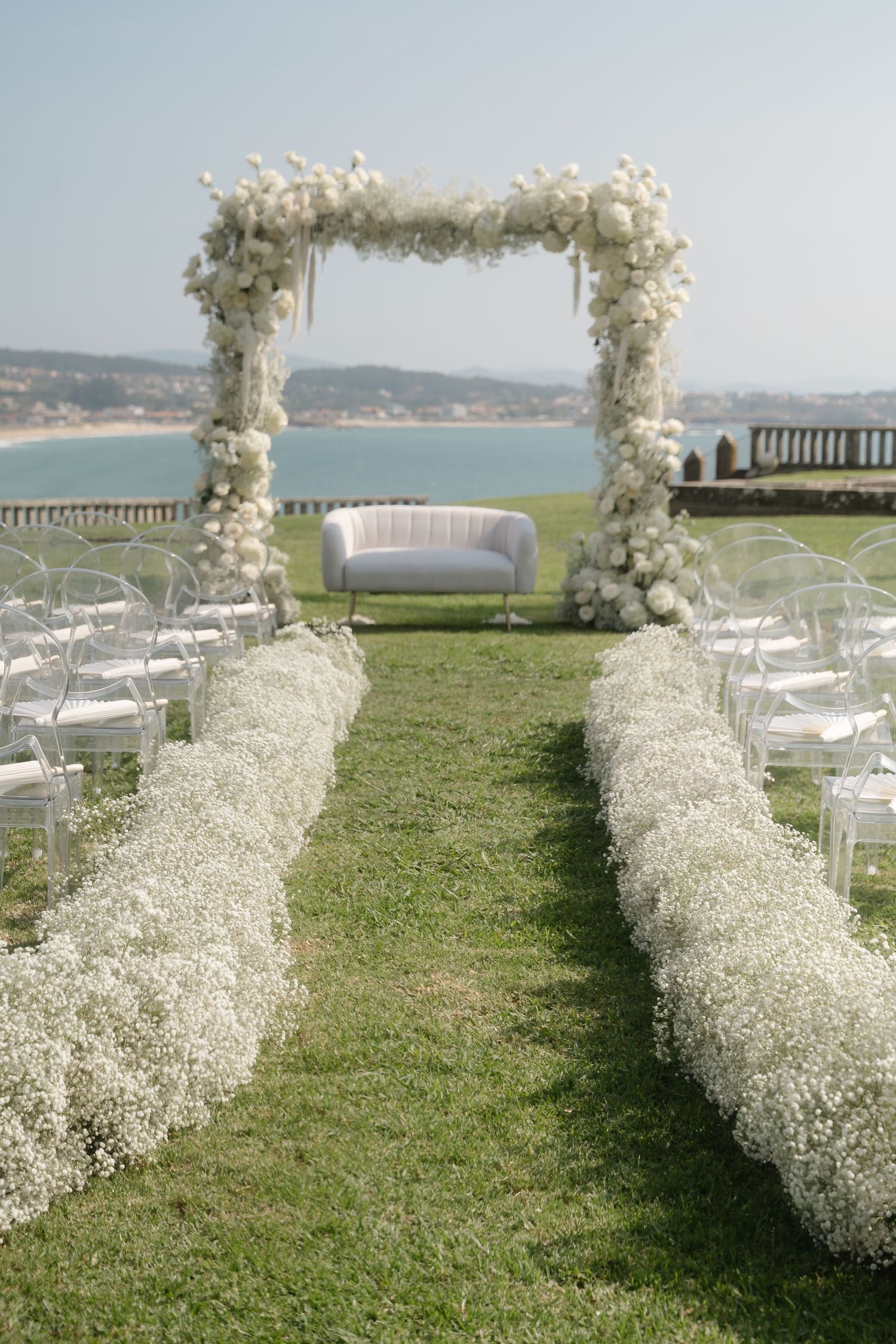 White floral arches and baby's breath line an outdoor wedding aisle overlooking a scenic coastal view.