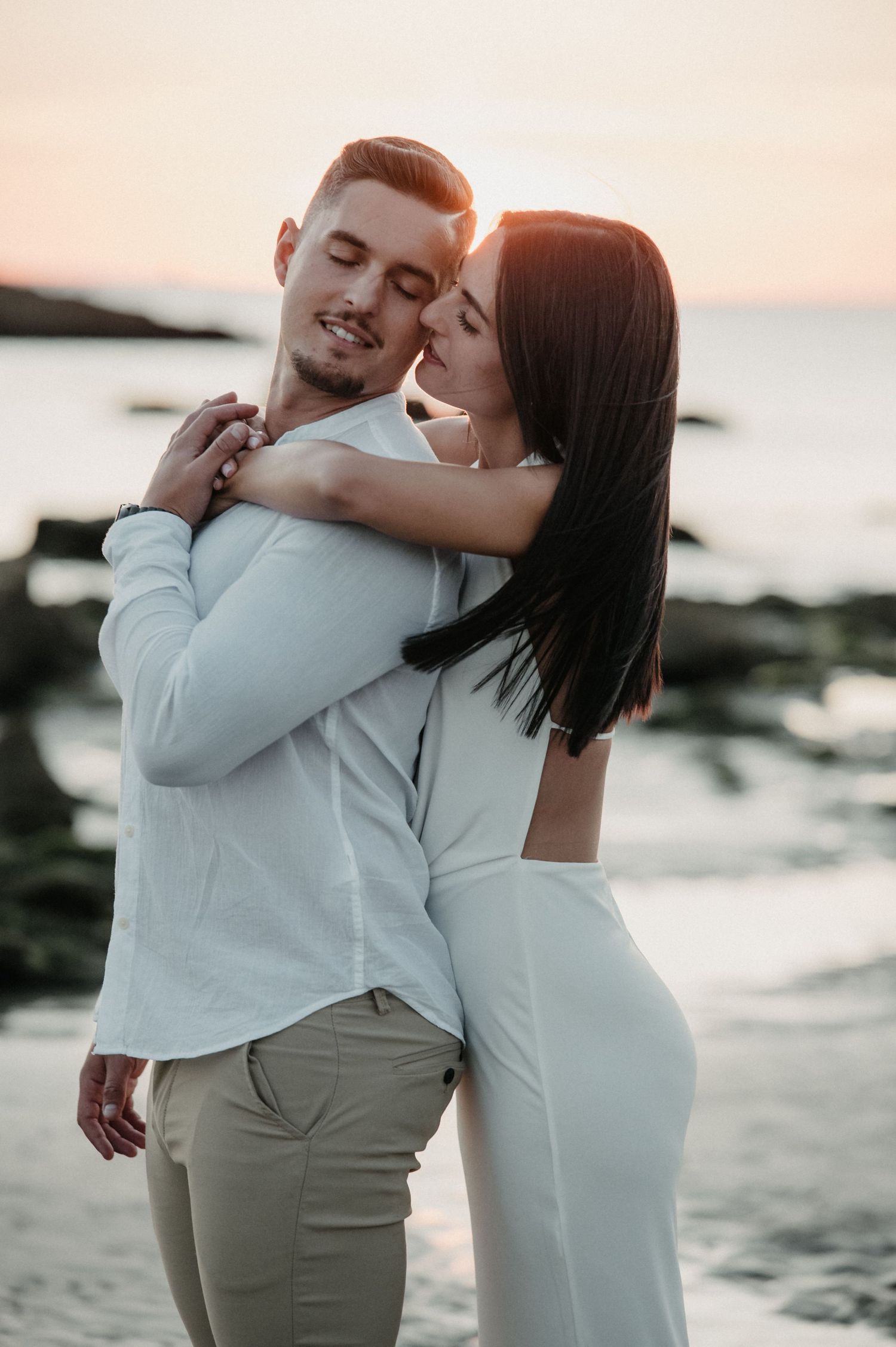 Intimate couple embracing on a rocky beach during golden hour with ocean in background.