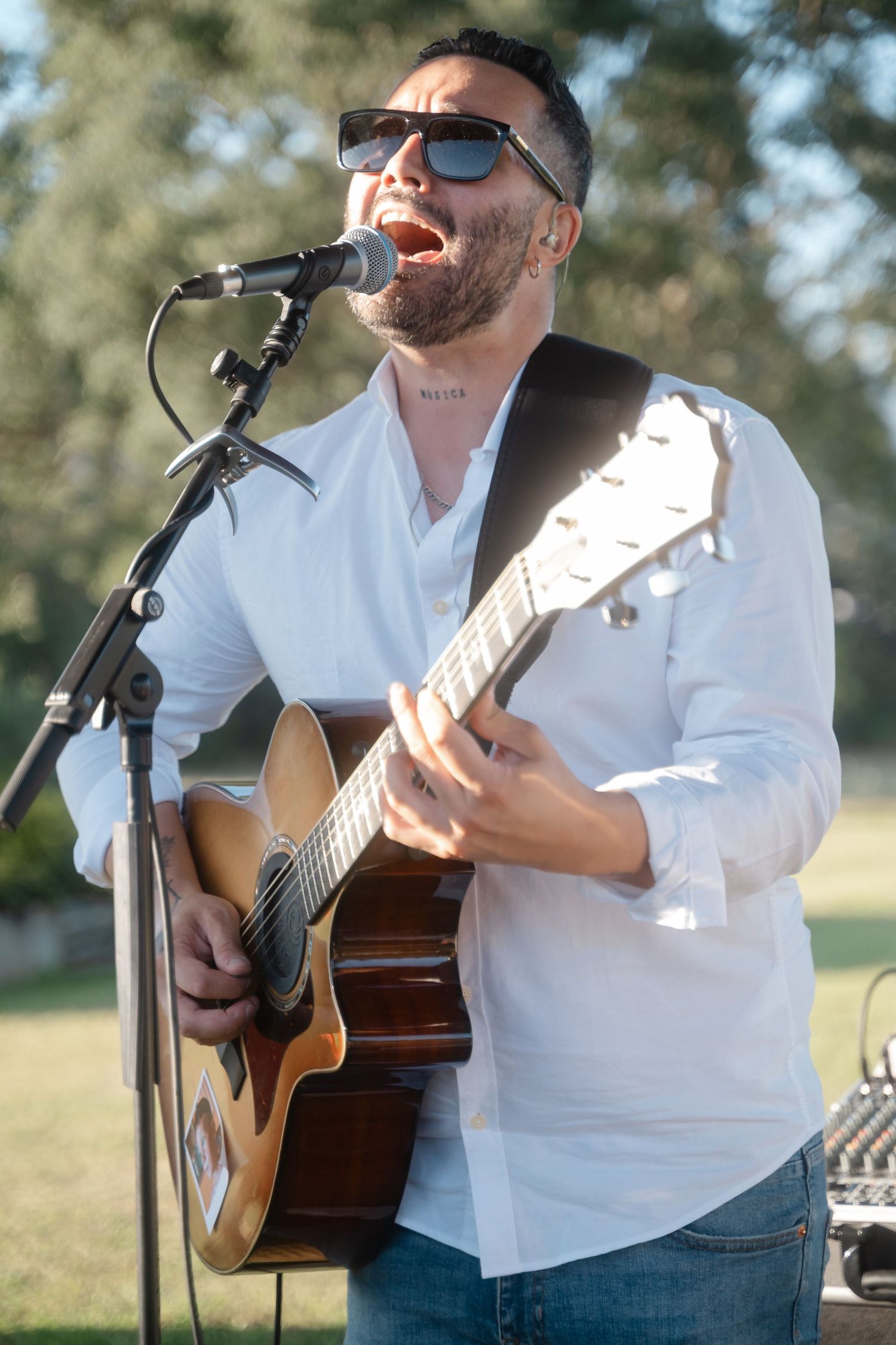 Musician in white shirt playing acoustic guitar at outdoor event with sunlit trees in background.