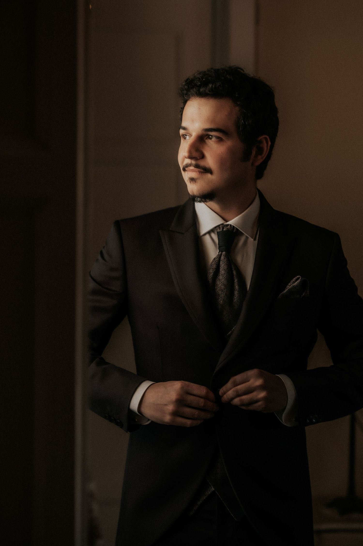 A person in a dark suit and tie stands in moody lighting against a shadowy wall, creating a dramatic portrait.