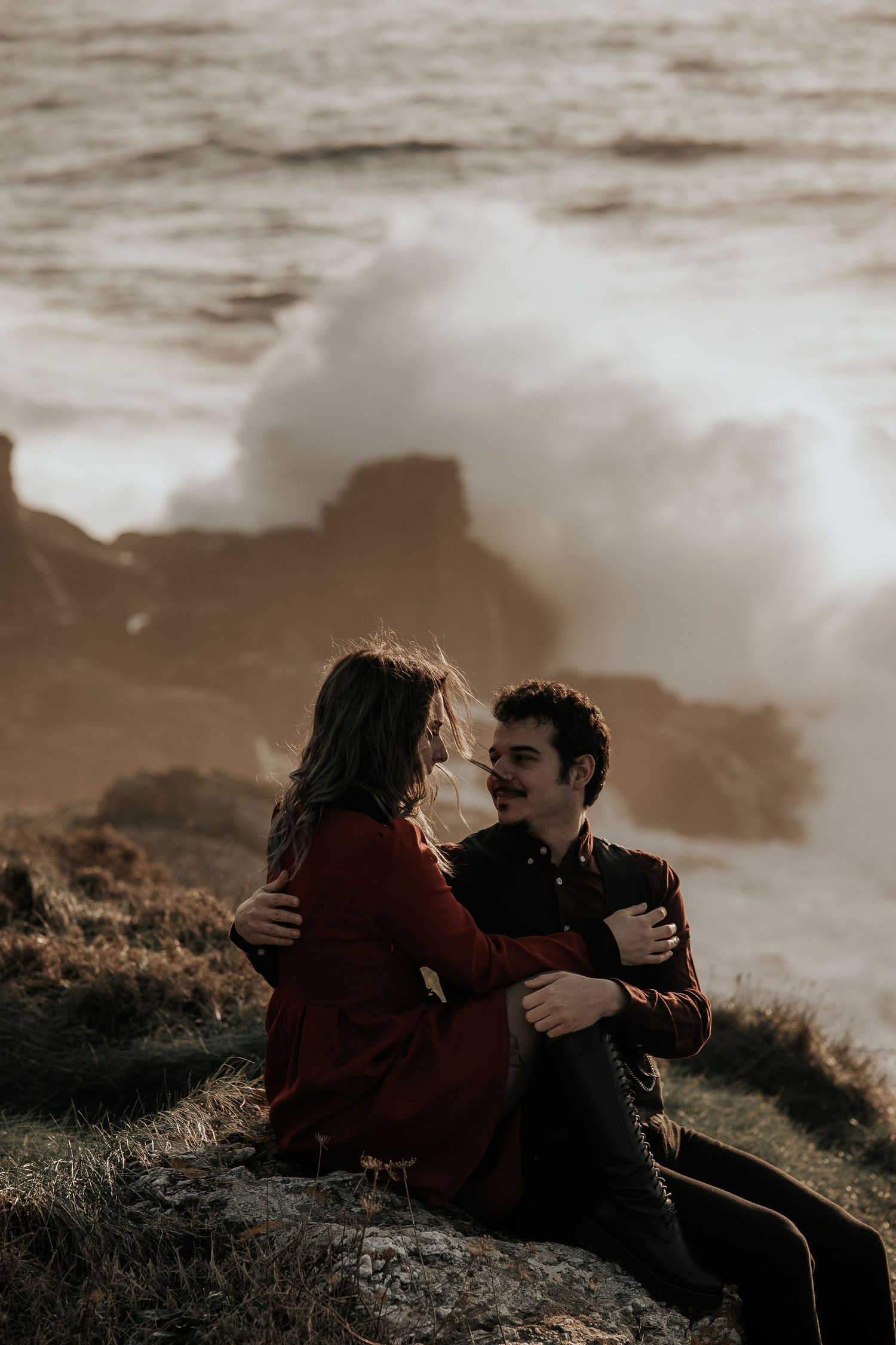 A couple sits together on a hillside during sunset with dramatic cloudy skies in the background.