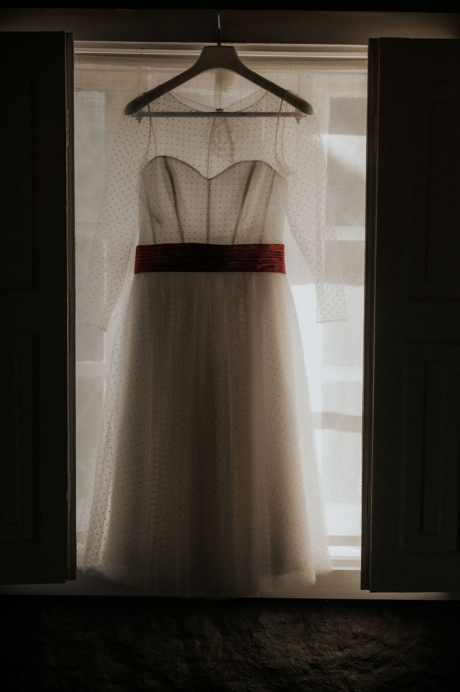 A white tulle dress with a red waistband hangs on a wooden hanger against a backlit window creating a moody silhouette.
