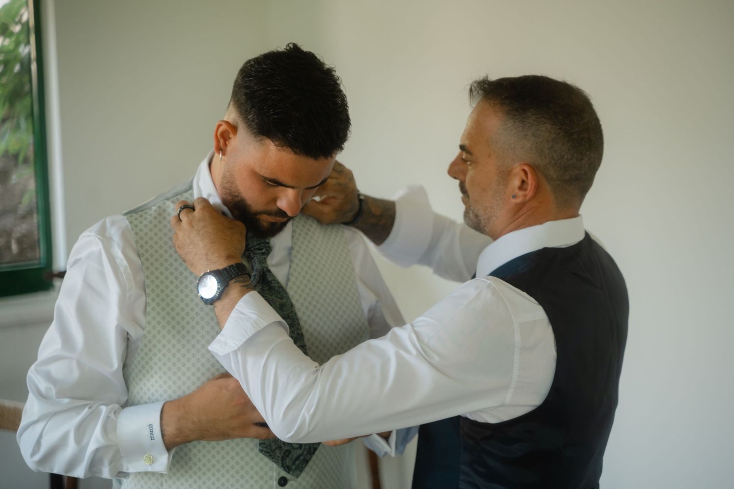 Two people in formal attire help each other get dressed, adjusting ties and vests before a special event.