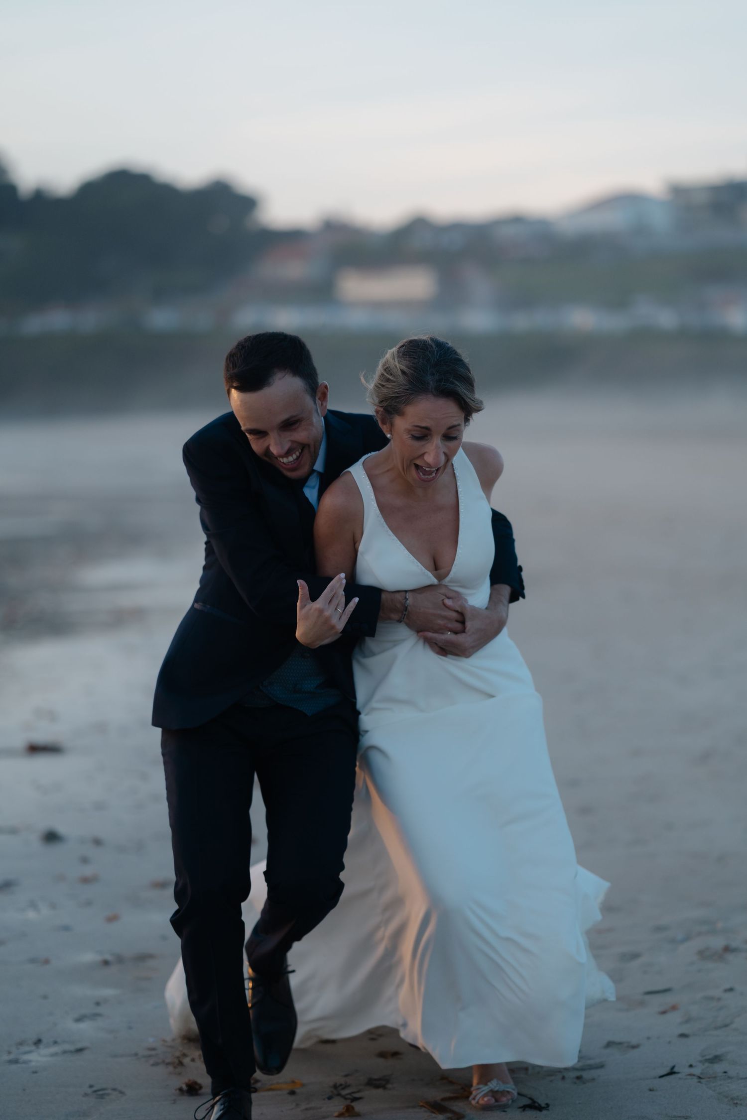 A couple in wedding attire laughs together while walking along a beach at dusk.