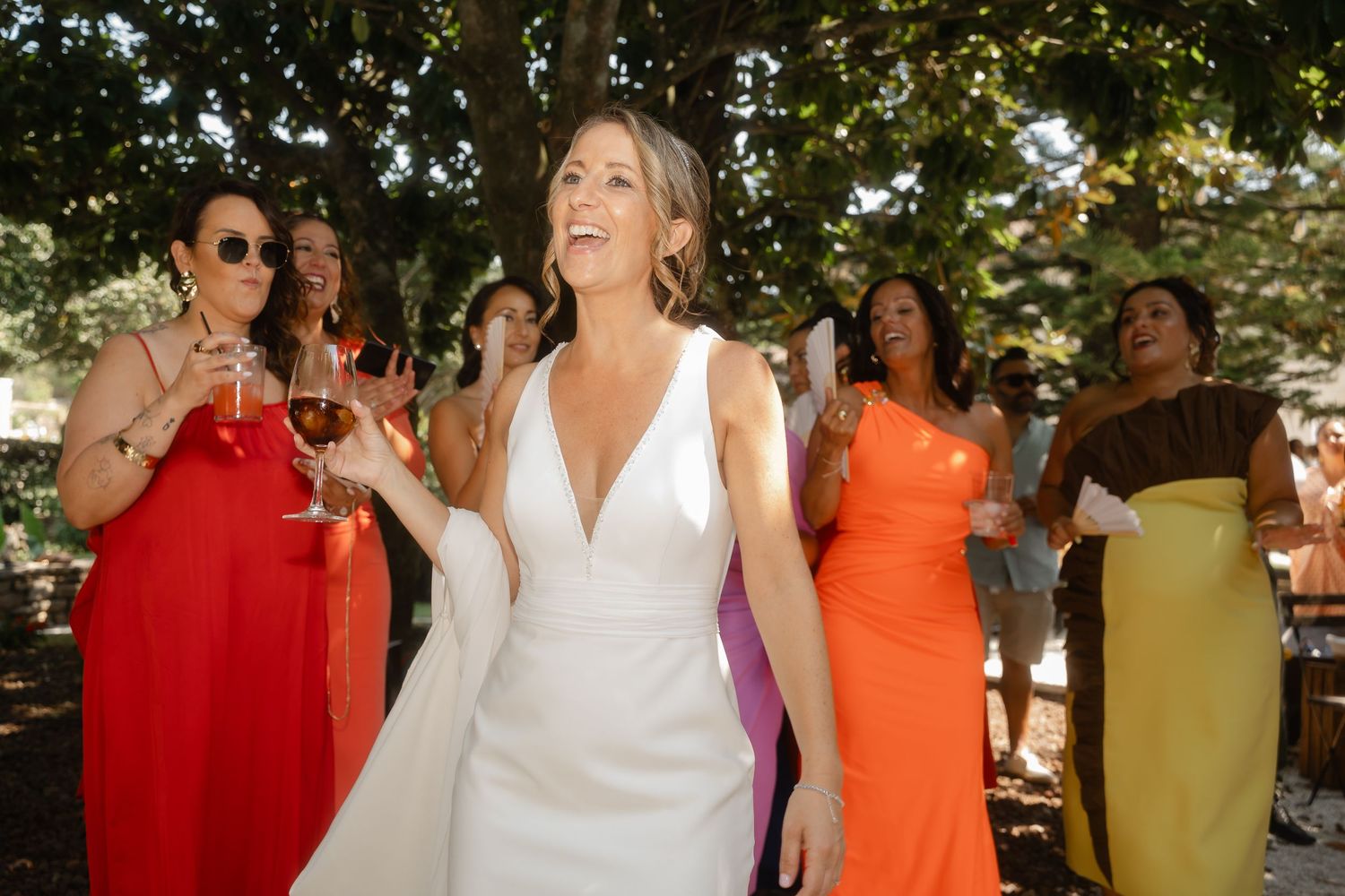 Wedding party members in vibrant orange, red, and white dresses pose together under trees.