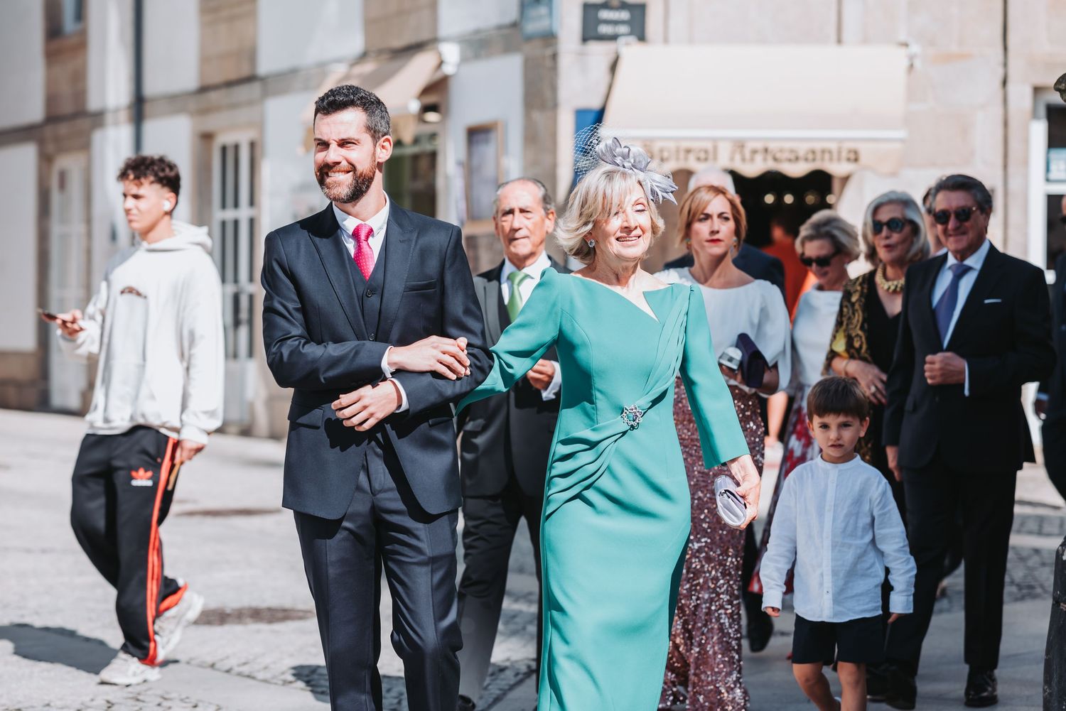 A group of wedding guests walk down a street with someone wearing a turquoise dress leading the procession.