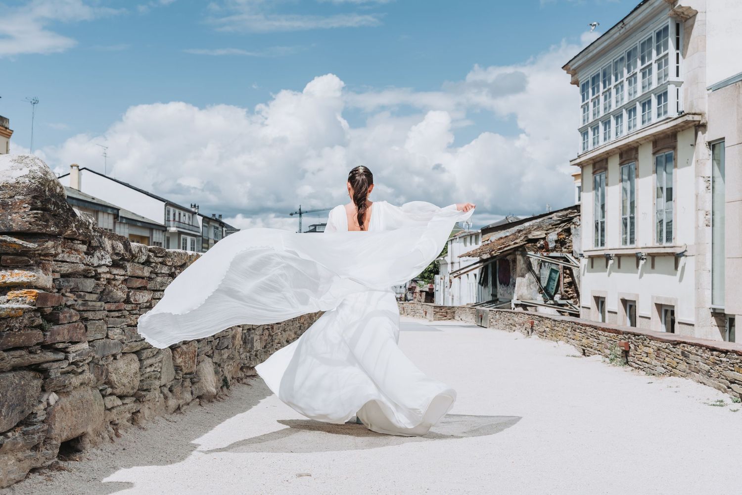 A flowing white wedding dress billows dramatically in the wind against a stone wall and blue sky backdrop.