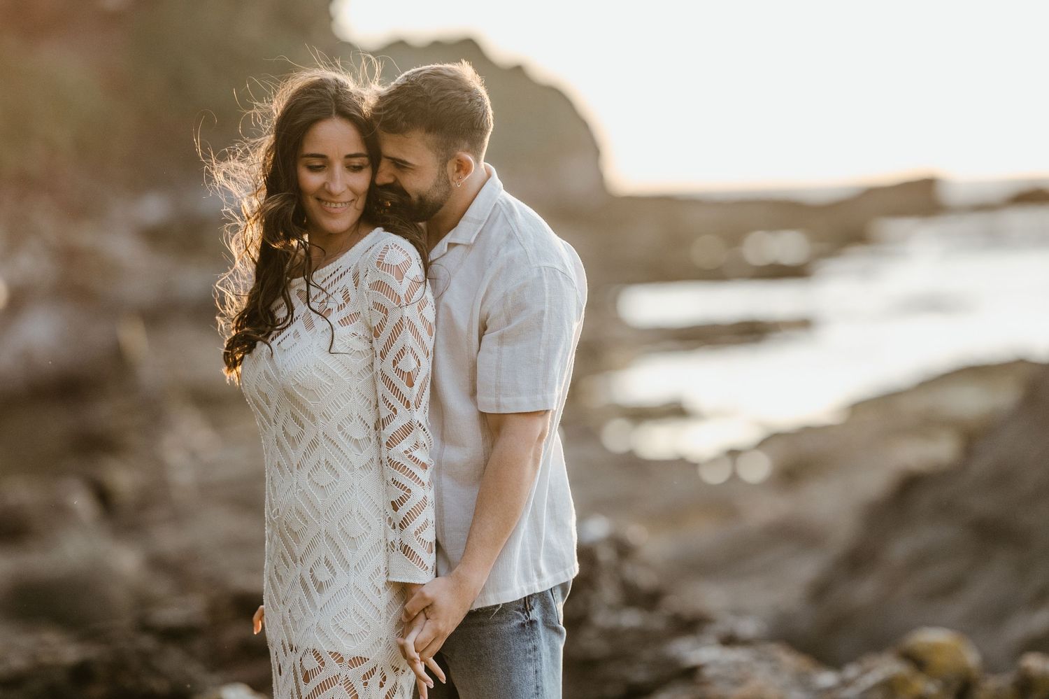 Couple in casual white outfits sharing tender moments against a coastal rocky landscape at golden hour.