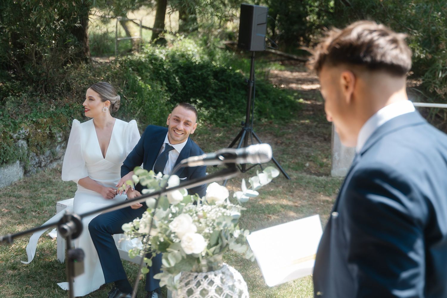 A couple sits together on a white bench in a garden setting during their wedding celebration.