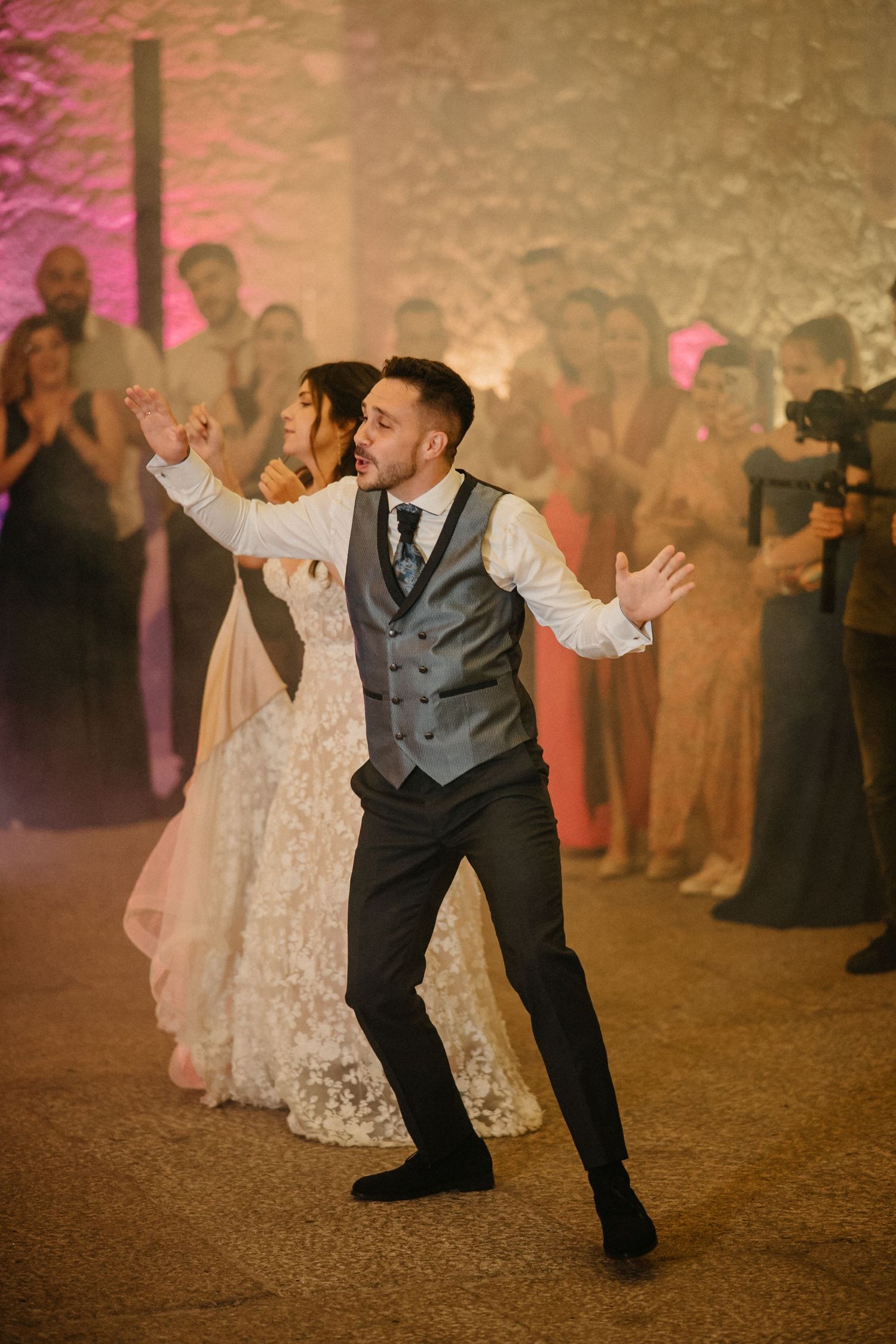 A couple performs an energetic dance move at their wedding reception with guests watching in the background.