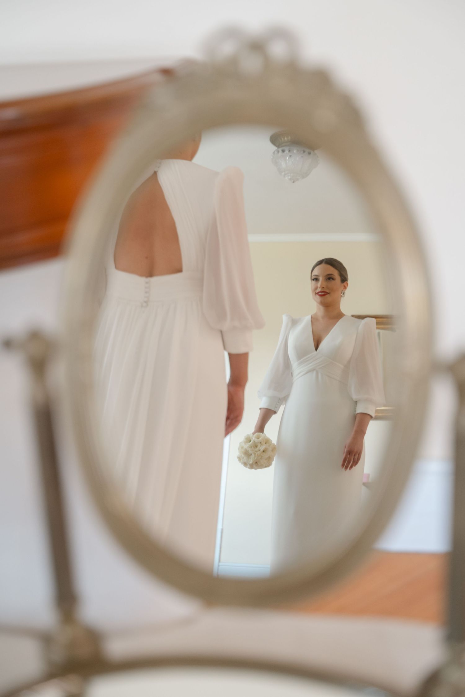 Elegant white wedding dress with long sleeves reflected in an ornate vintage mirror.