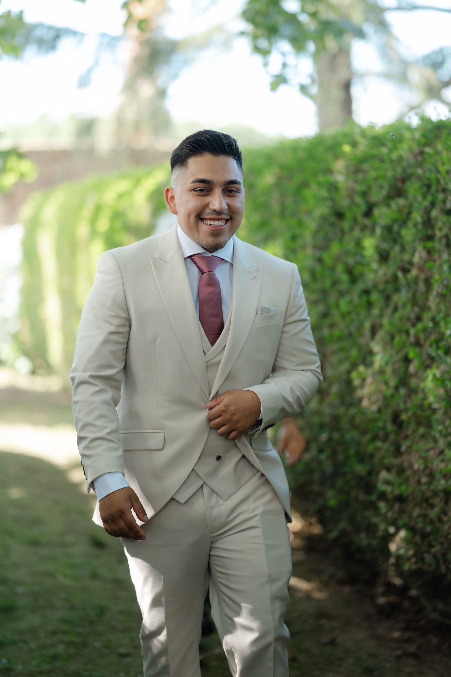 Groom in a light beige suit and burgundy tie smiles joyfully in an outdoor garden setting.