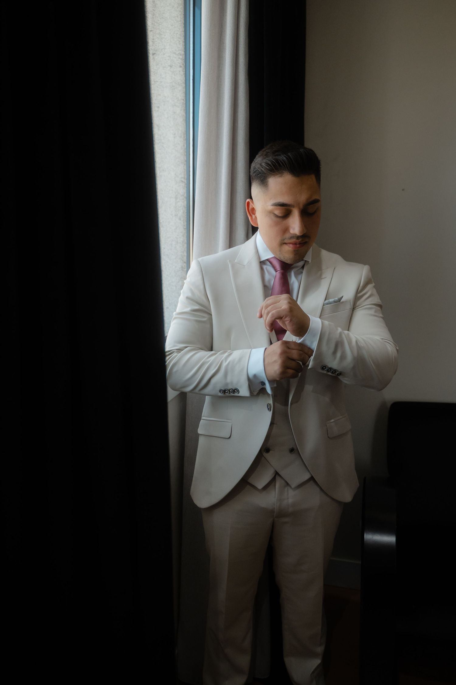A person in a crisp white suit and pink tie adjusts their cuff while standing by a window in moody lighting.