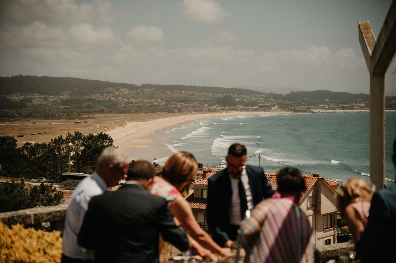 Panoramic coastal view showing a sandy beach with waves crashing along the shoreline and hills in the background.