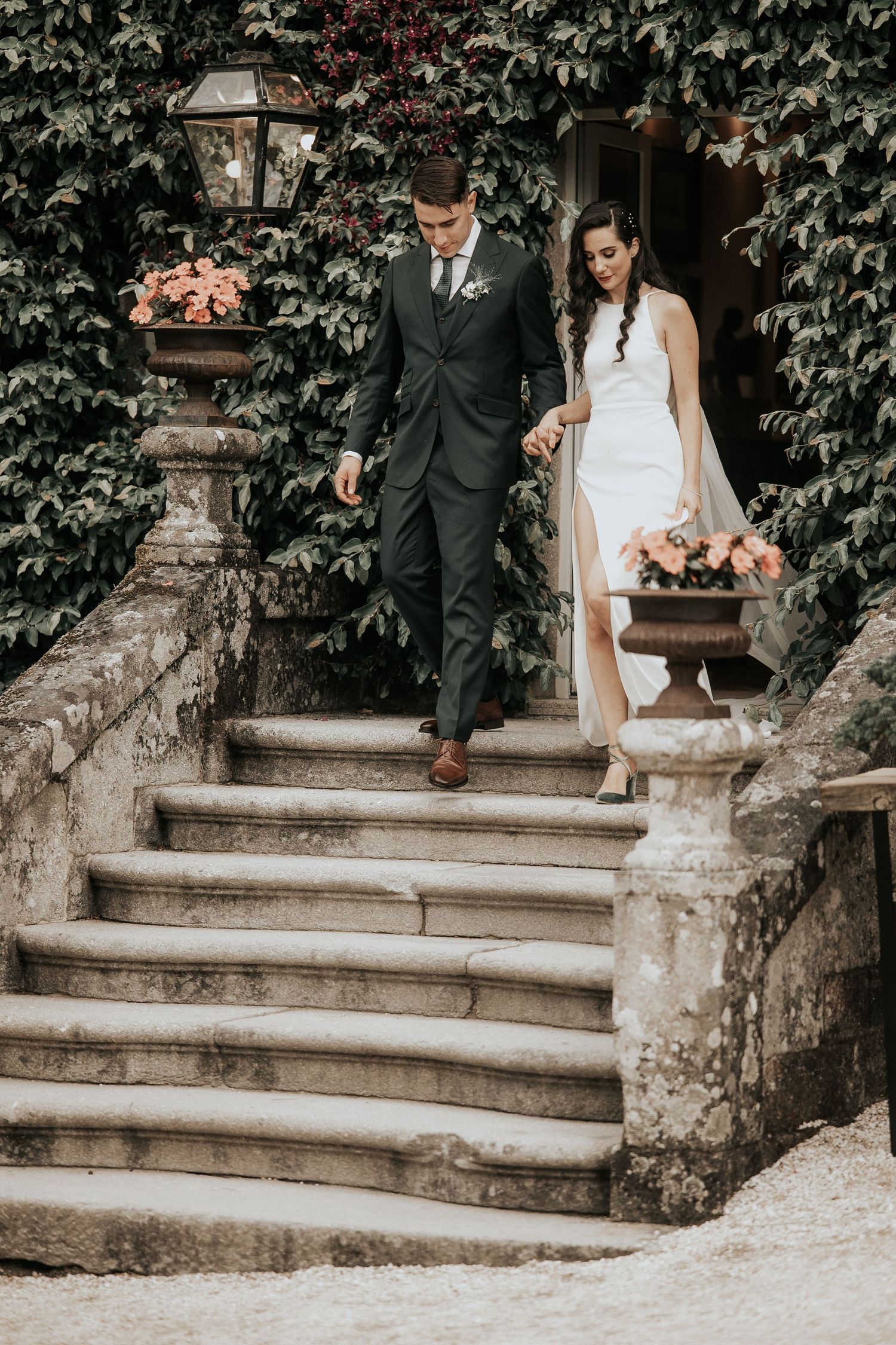 A couple in formal wedding attire descends stone steps surrounded by ivy-covered walls.