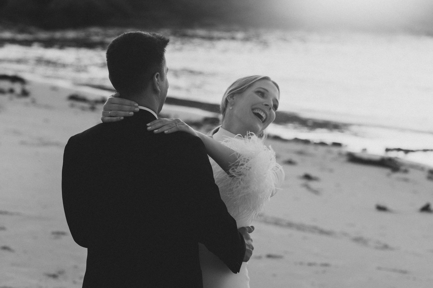 Black and white photo of newlyweds dancing together on a beach at sunset.