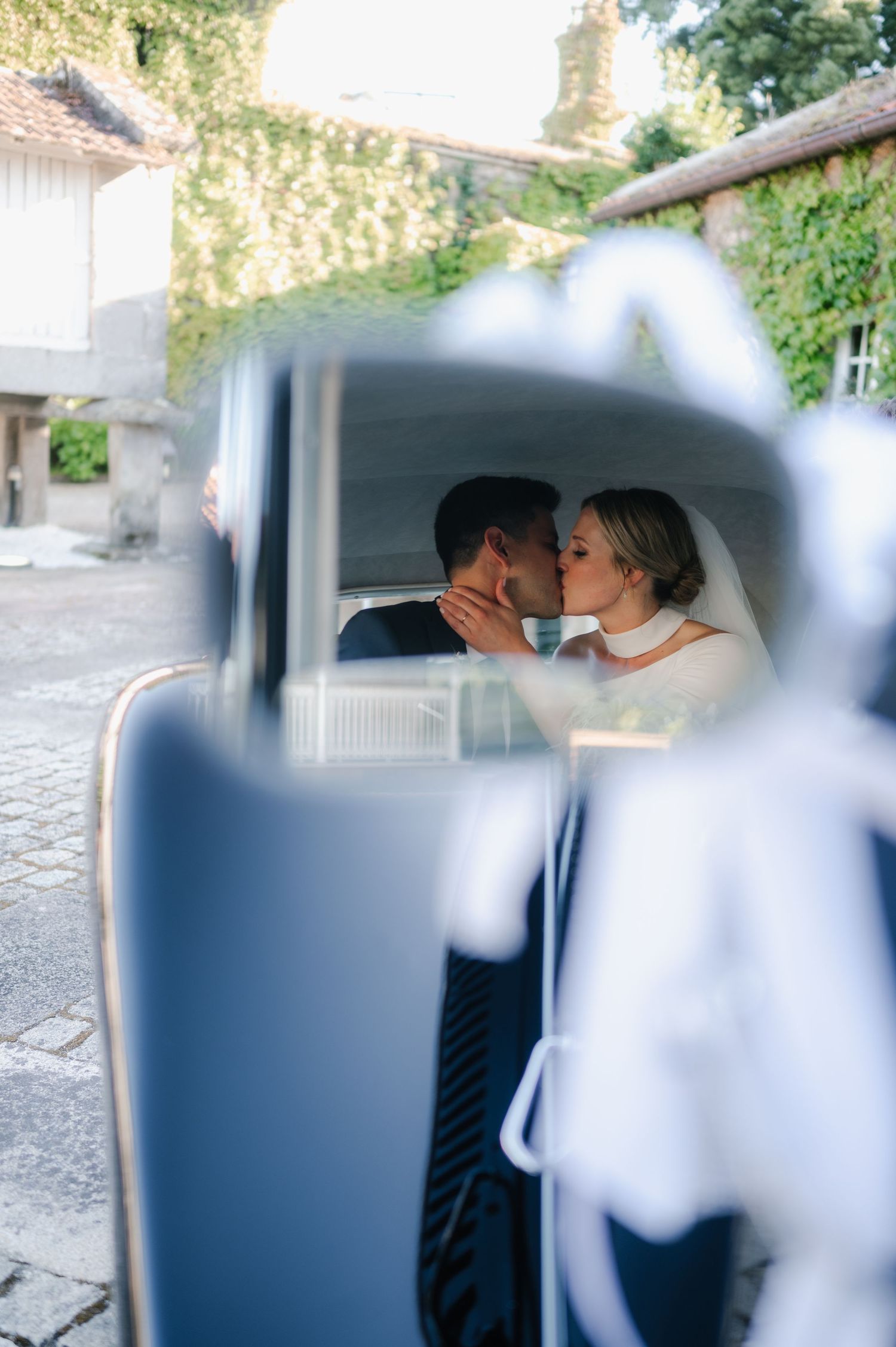 A romantic moment captured through a car window shows a bride in white sharing a tender kiss on their wedding day.