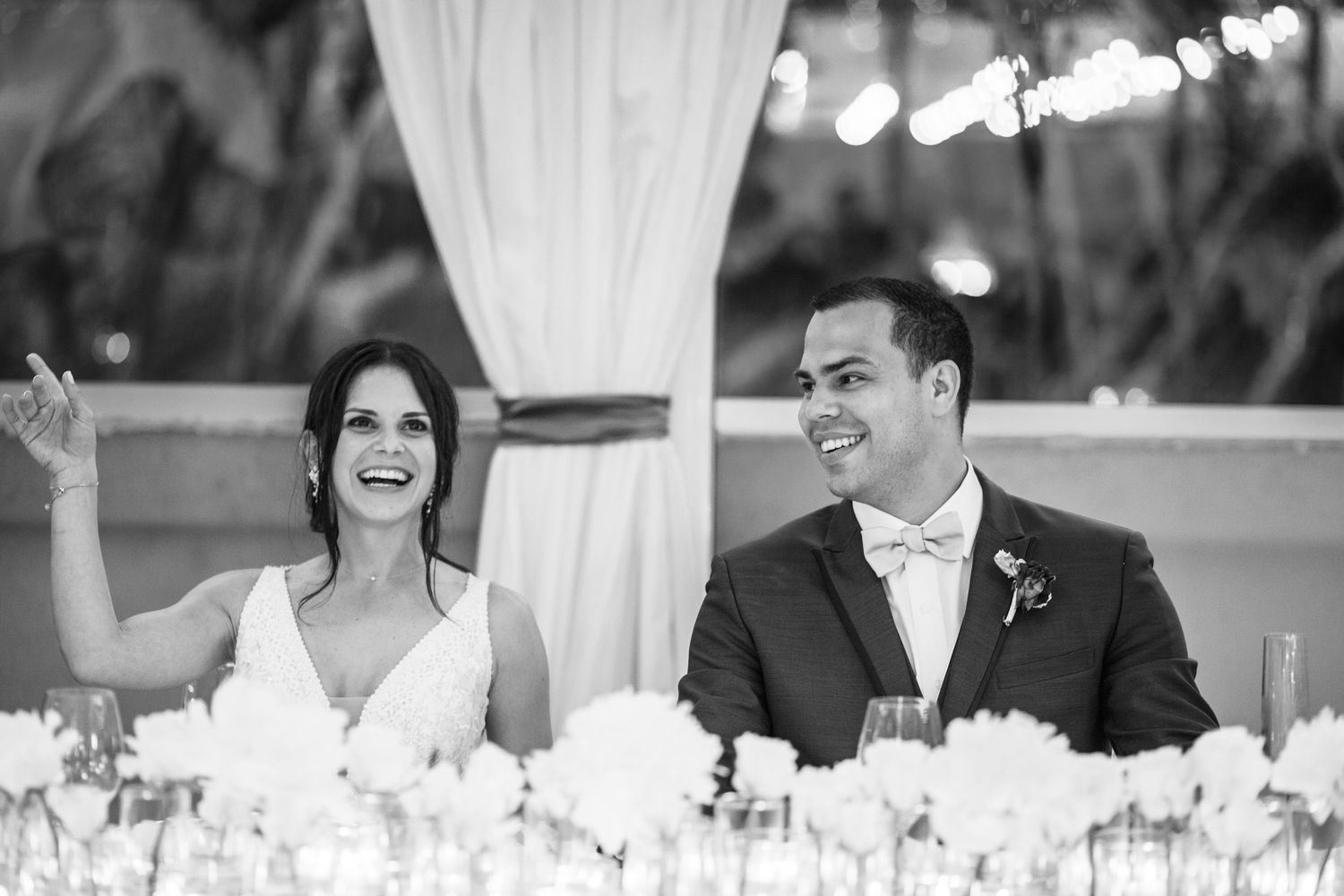 Wedding guests seated at outdoor reception tables with white linens and glowing string lights overhead during sunset.