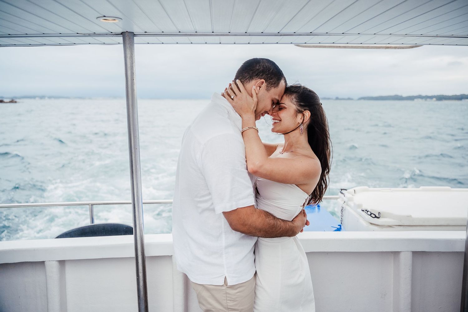 A couple embraces romantically on a boat deck with ocean views in the background.
