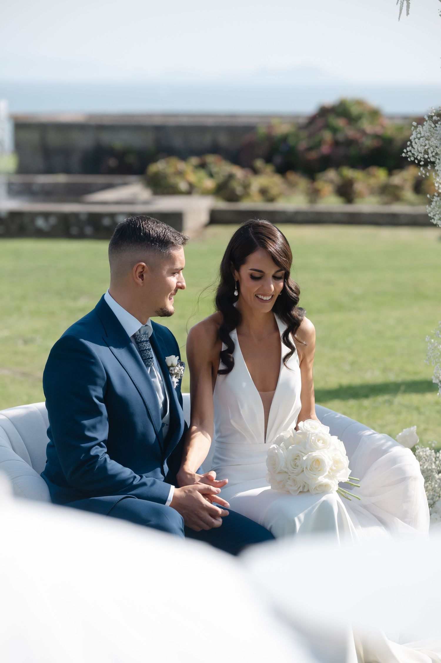 A romantic wedding couple sits together on grass overlooking a scenic coastal view on their special day.