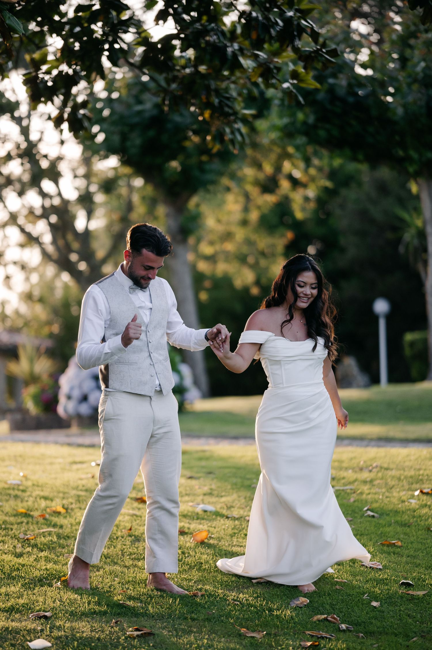 A couple in wedding attire dances playfully on a grassy hill during sunset in a park setting.