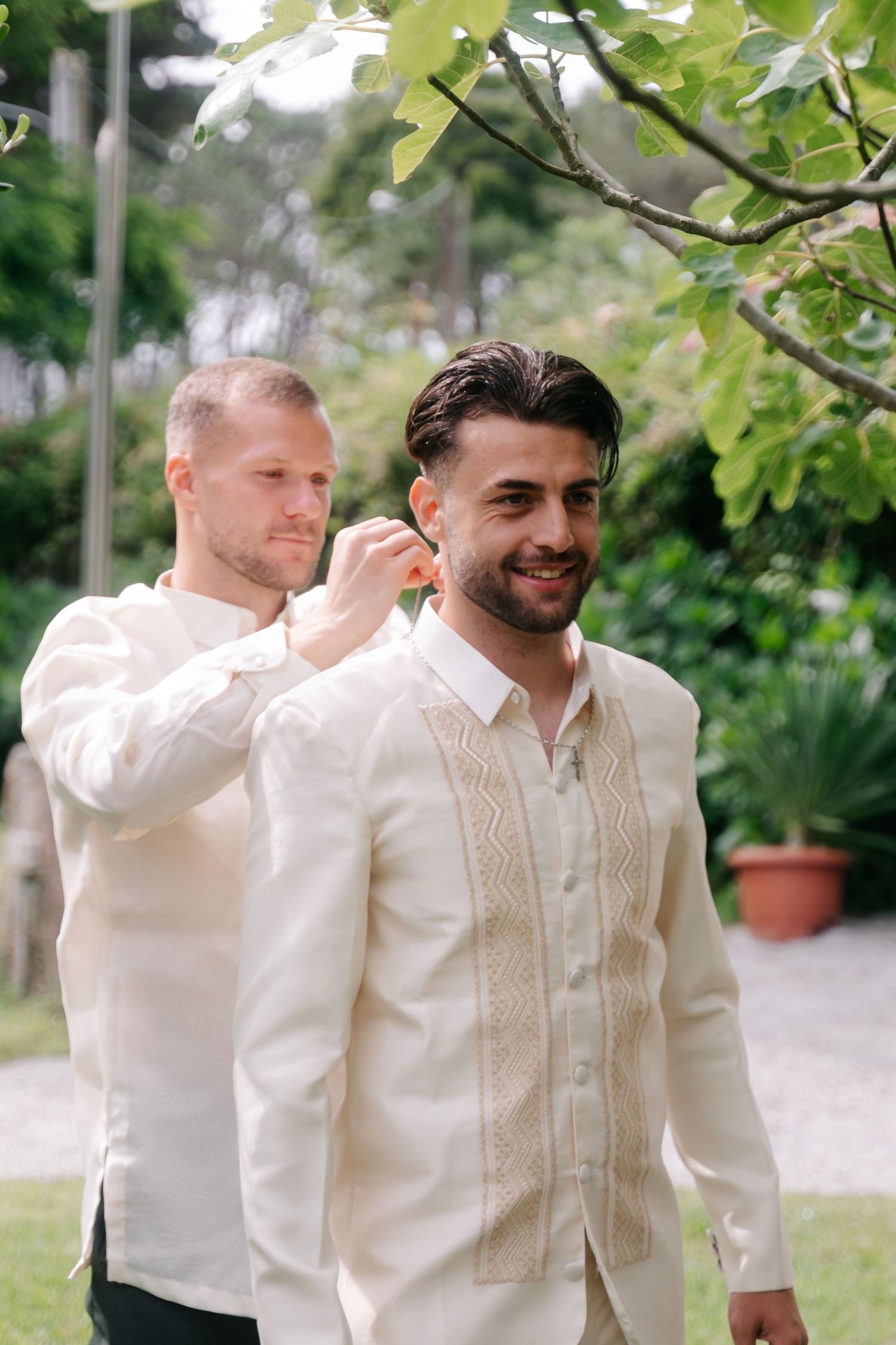 Two people in traditional white embroidered attire share a lighthearted moment outdoors in a garden setting.