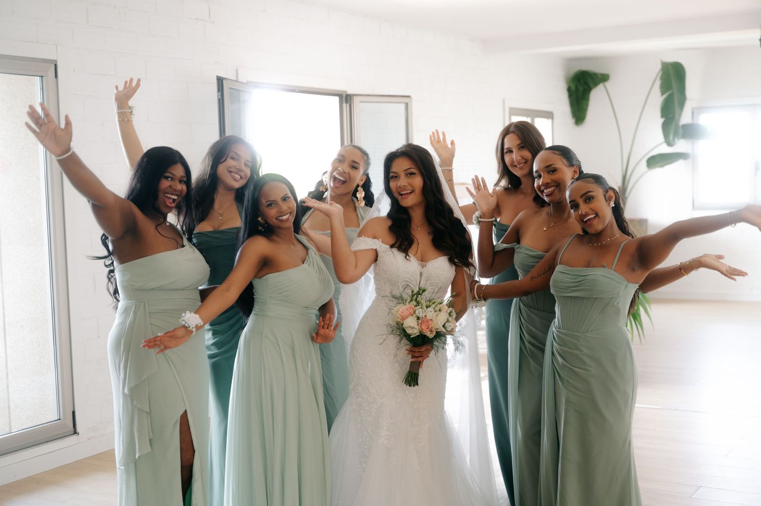 A group in matching sage green bridesmaid dresses pose playfully in a bright white room.