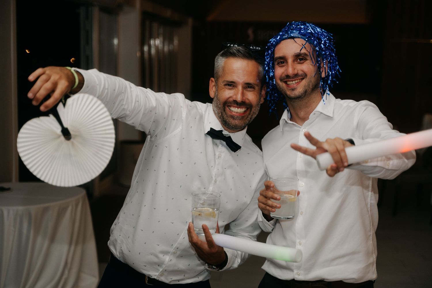 Two wedding guests in white attire pose with paper fans and drinks during reception celebrations.