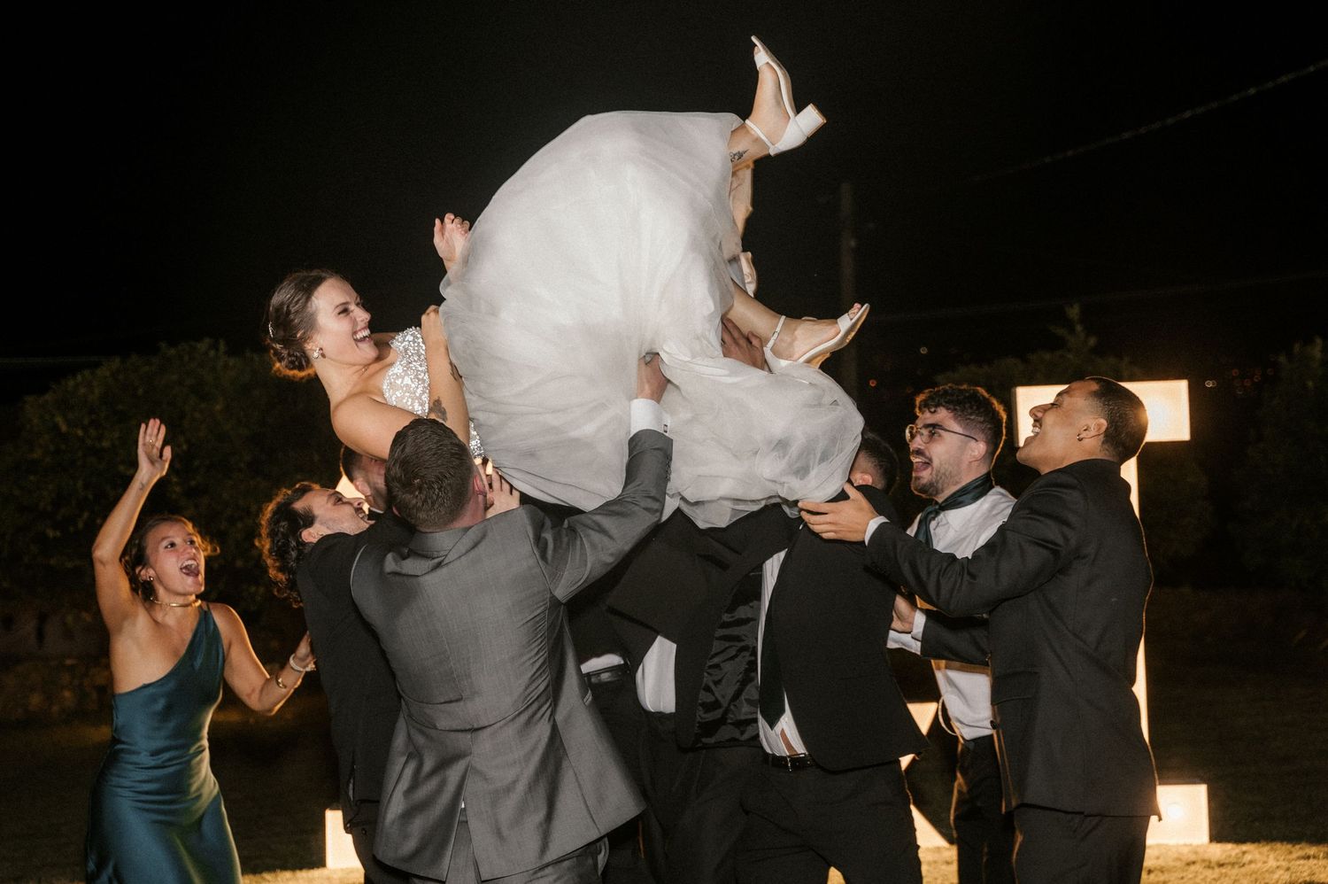 Wedding party lifts bride in her white dress during an energetic outdoor reception celebration.