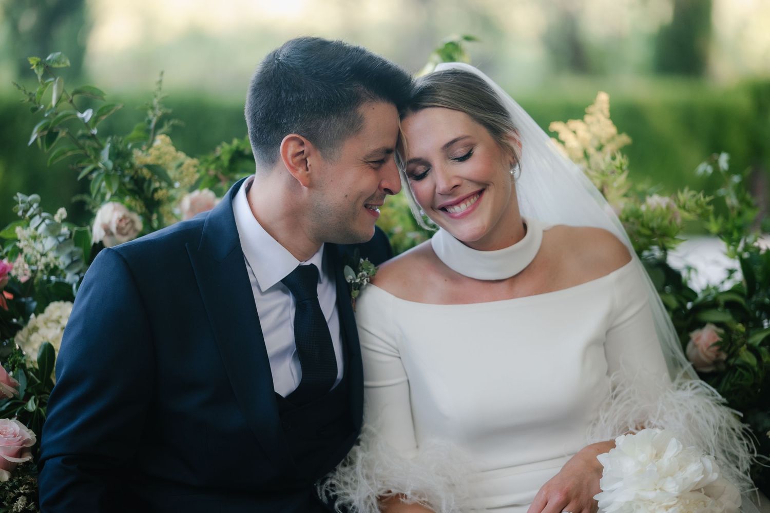 A romantic wedding couple embraces outdoors surrounded by lush greenery and natural light.
