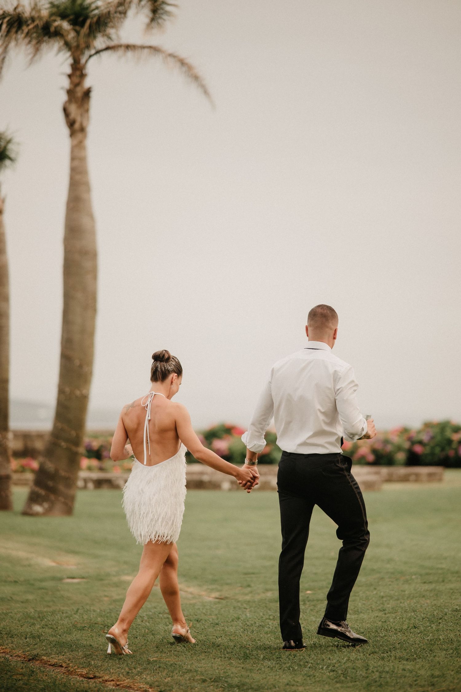 A couple dances playfully on a grassy lawn lined with palm trees during what appears to be a wedding celebration.