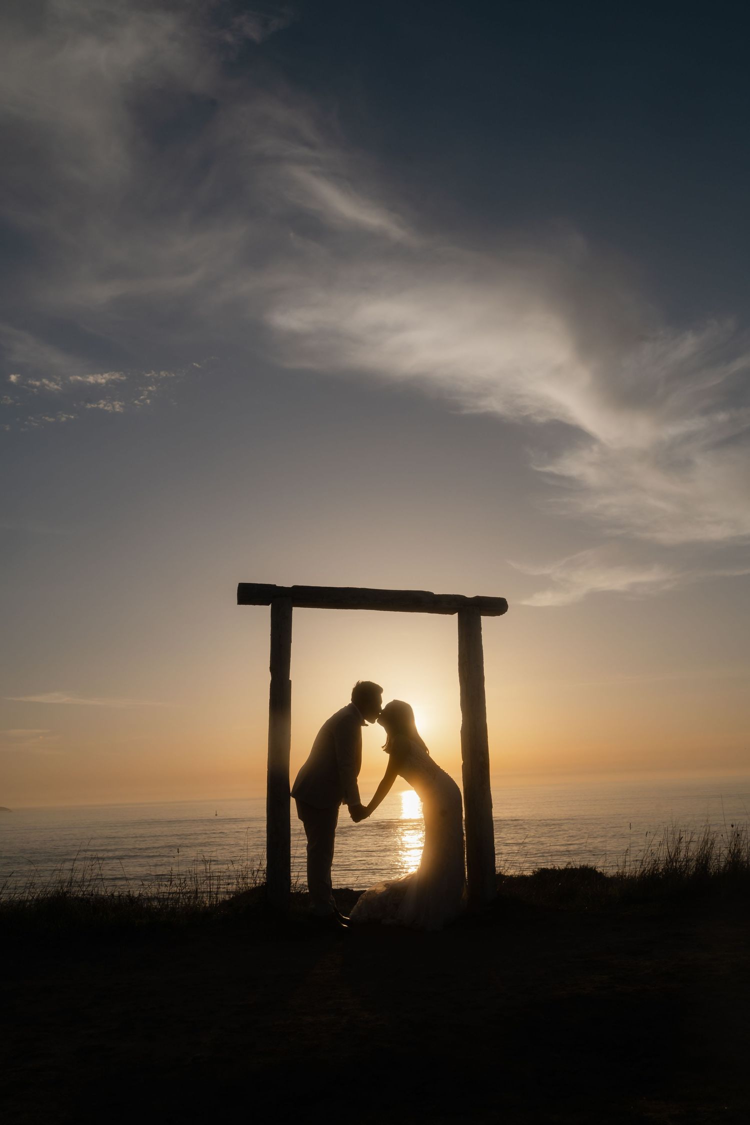 Silhouetted figures create heart shape against wooden archway during dramatic sunset over ocean horizon.