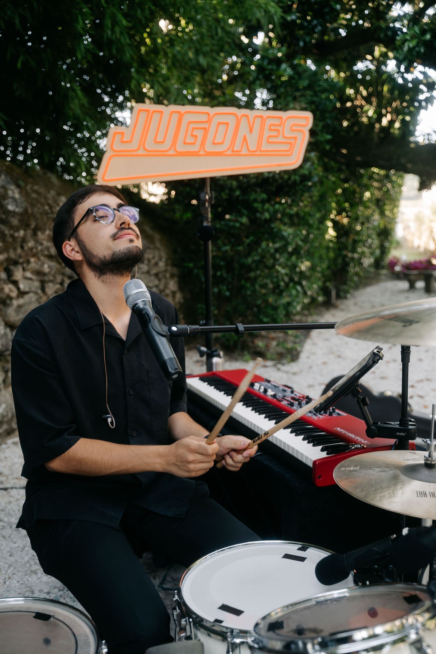 Musician performs behind drum kit with red keyboard and Jugones sign at outdoor venue.