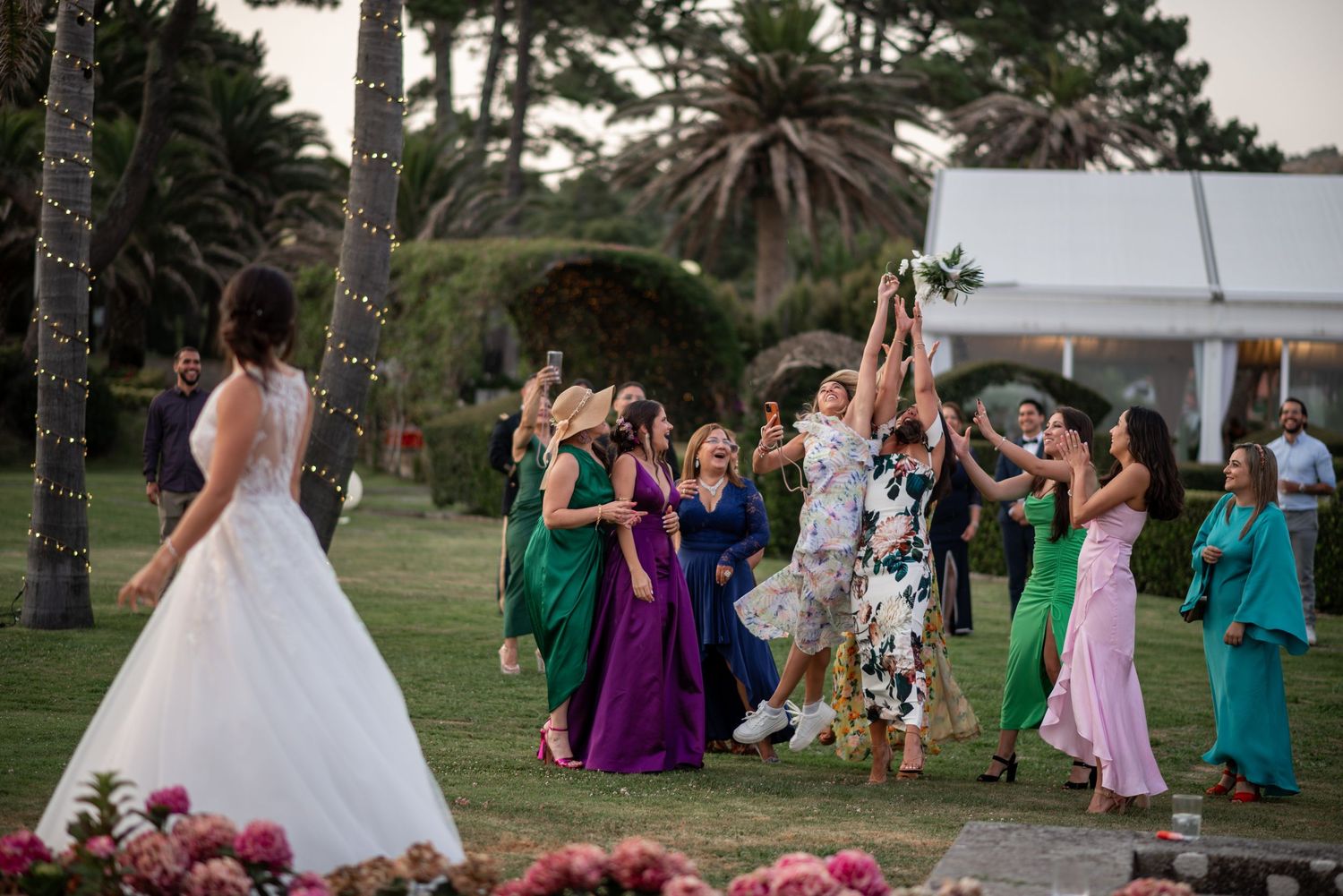 Outdoor wedding celebration with guests in colorful dresses gathered under trees.