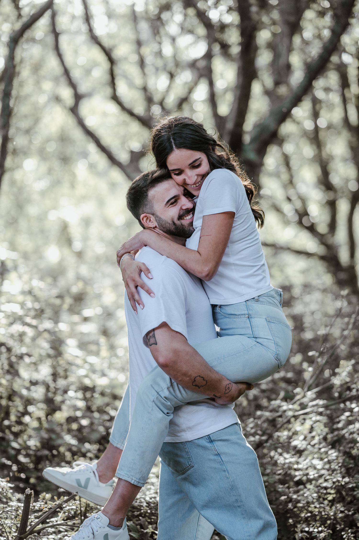 A joyful couple in casual white and denim outfits sharing a playful embrace among trees in natural lighting.