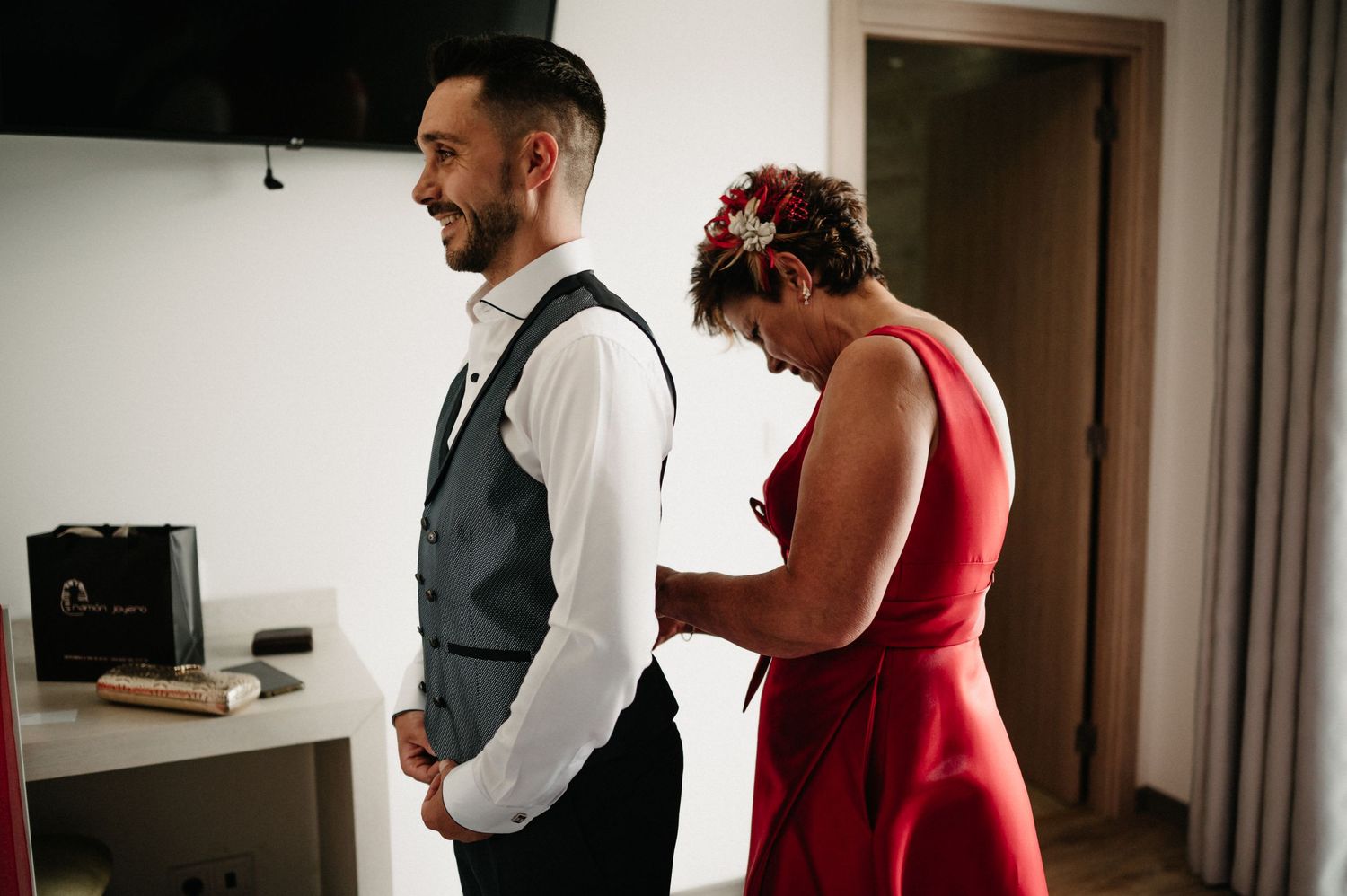 A couple in formal attire, with a red dress and grey vest, share a tender moment in an interior hallway.