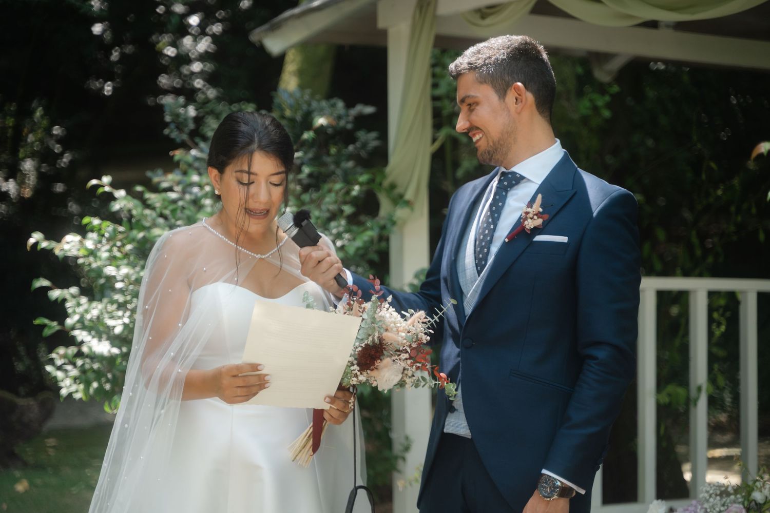 Couple participates in outdoor wedding ceremony under white gazebo with natural lighting.