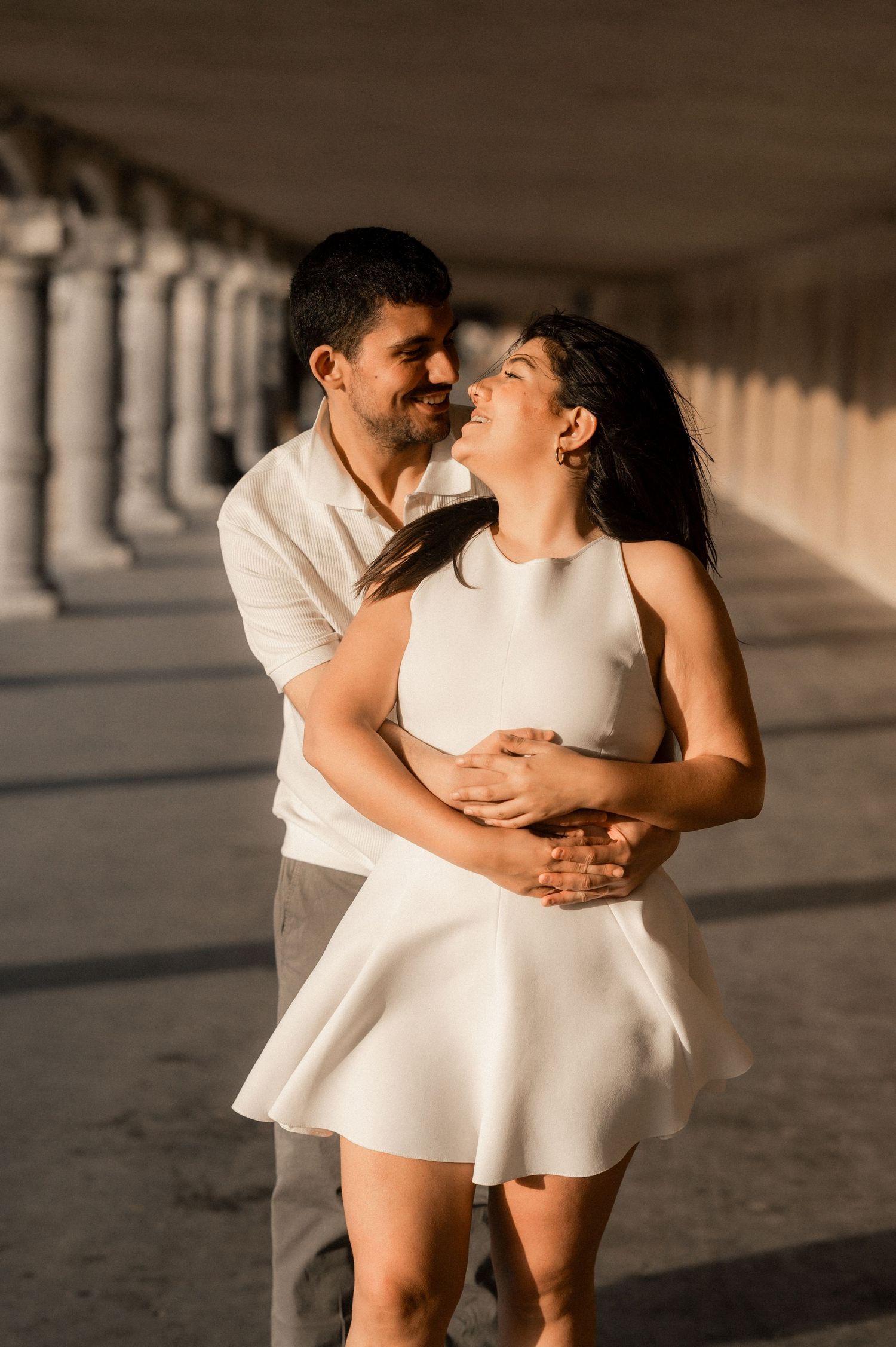 Couple in white outfits dance and embrace under architectural columns in warm sunlight.