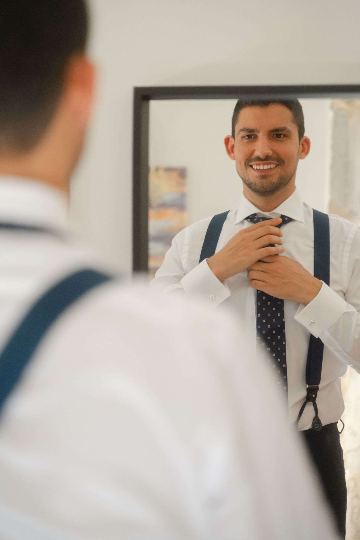 A person in a white dress shirt and navy tie adjusts their collar while looking in a mirror.