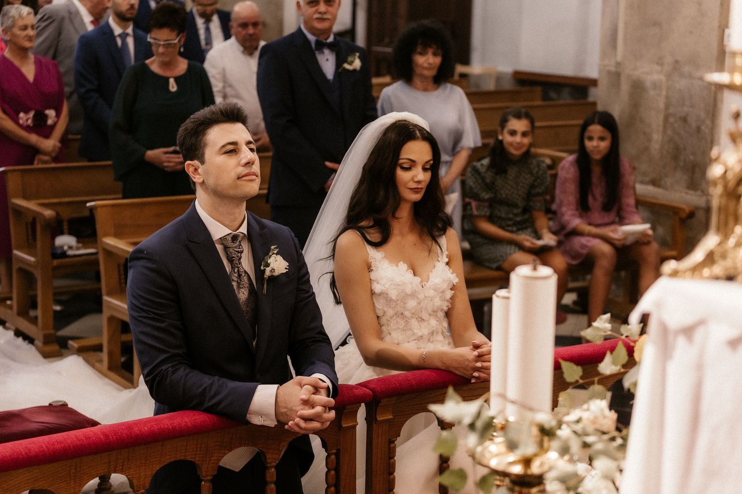 A couple sits in church pews during a traditional wedding ceremony surrounded by guests and candlelight.