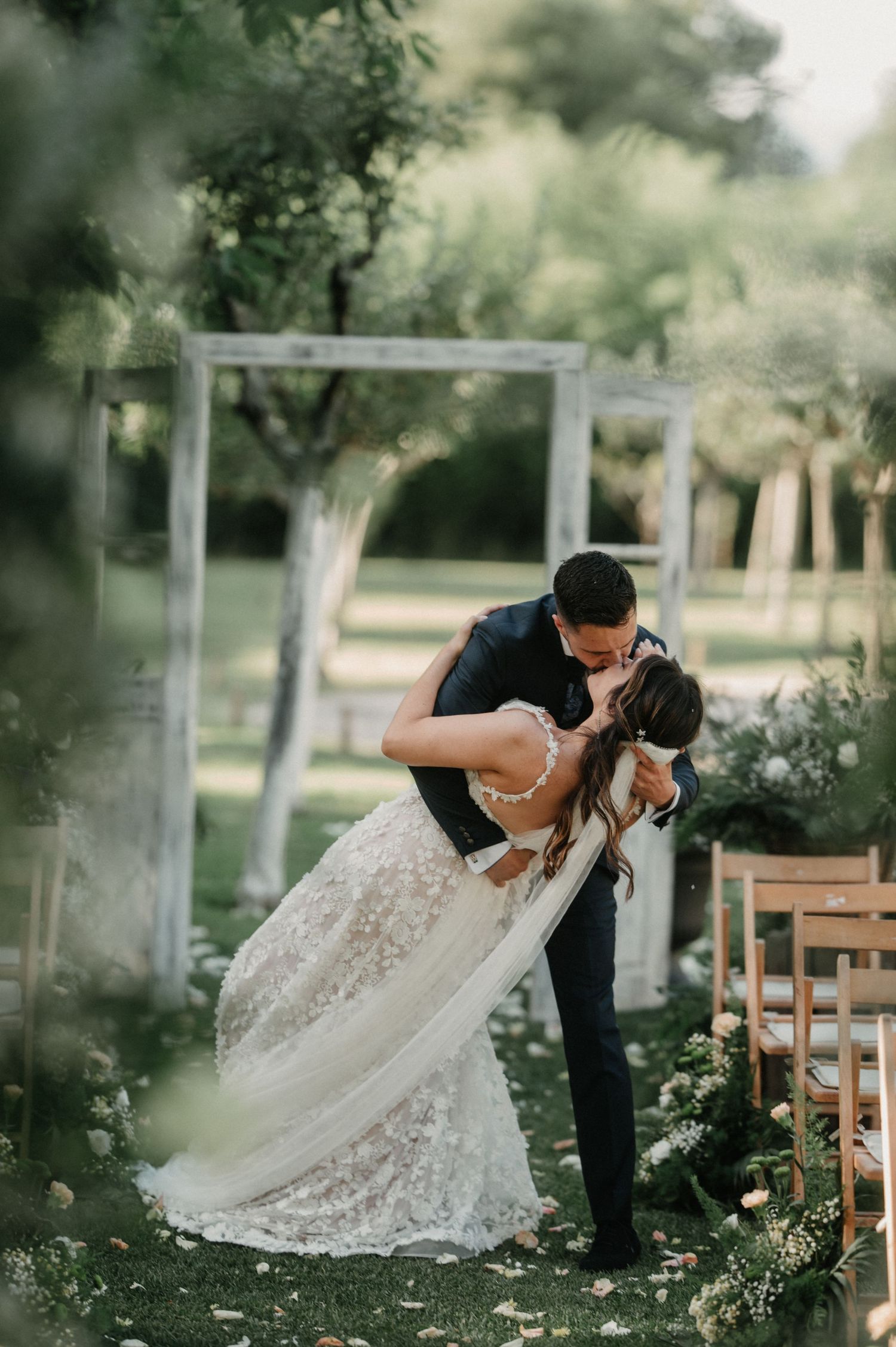 A romantic wedding couple shares intimate moments in a garden setting with wooden chairs and dreamy lighting.